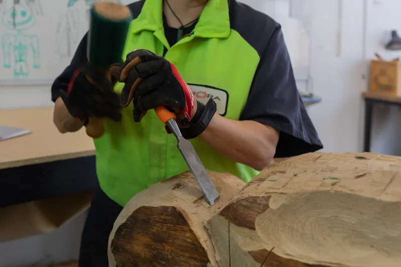 A person in a bright green shirt and gloves uses a mallet and chisel to carve a large piece of wood in a workshop.