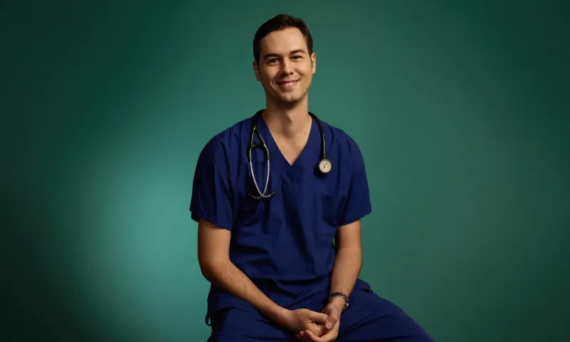 A photo of a young, male doctor wearing scrubs and a stethoscope. He is smiling.