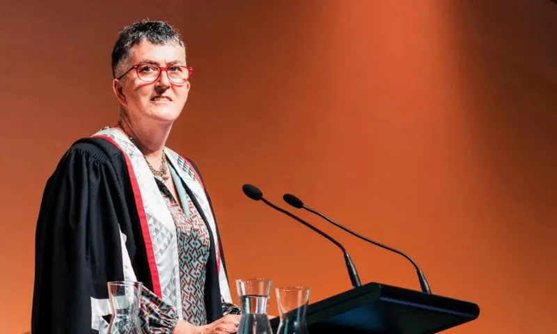A photograph of a woman with short hair standing at a lectern with an academic gown on. She looks as if she is looking over the audience, about to speak.