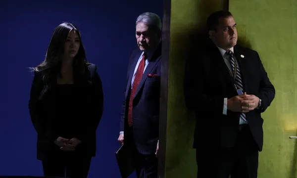Three people in business attire stand indoors, partially separated by a wall; two are in shadow on the left, and one stands in light on the right, all with serious expressions.
