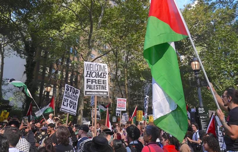 A large crowd of protesters gathers outdoors, holding Palestinian flags and signs with messages such as "Welcome Home Free Palestine" and "Stop Killing Let Gaza Live." Trees and buildings are visible in the background.