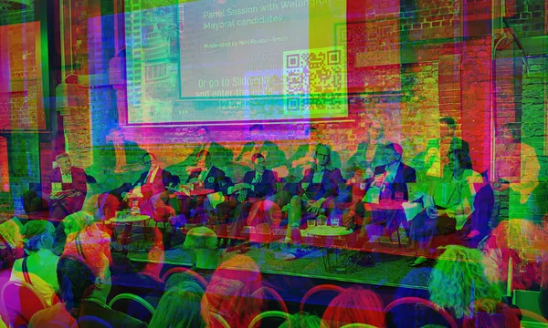 A panel of speakers sits on stage in front of an audience, with a large screen displaying event information and a QR code on a brick wall behind them. The event appears to be a discussion or forum.