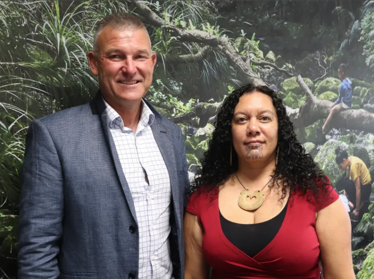 A man in a blue blazer and a woman in a red top with a traditional Māori necklace and chin tattoo stand together, smiling, in front of a lush forest background image.