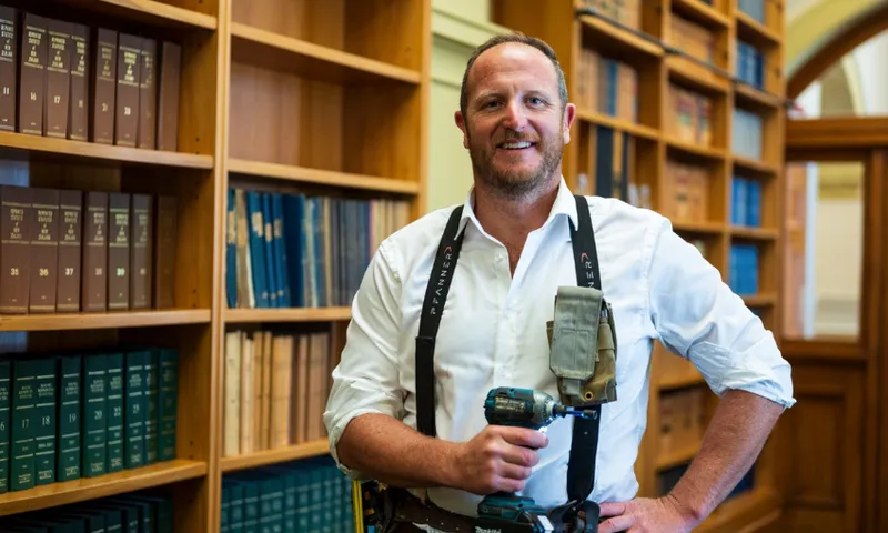 Cameron Luxton stands in a hallway in parliament wearing a tool belt and holding a drill.