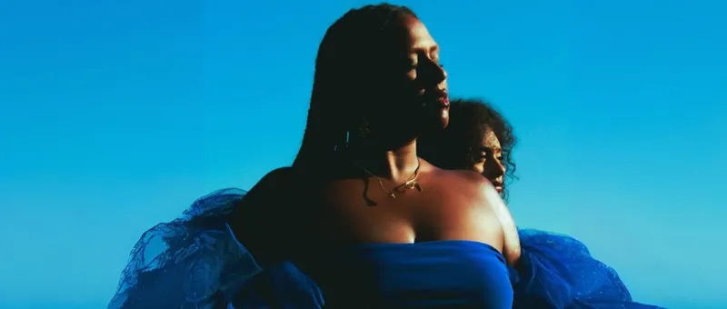 A cropped photo of singer Deva Mahal who is a dark-skinned woman. She is wearing a blue dress and there is a bright blue sky behind her.