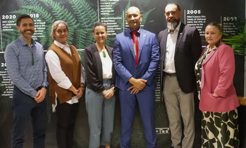 Six people stand in a row, dressed in business or smart casual attire, in front of a background featuring green ferns and text about historical events. They appear to be posing for a formal group photo.