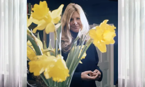 A young Jilly Cooper stands in her home with a bunch of daffodils in front of her.