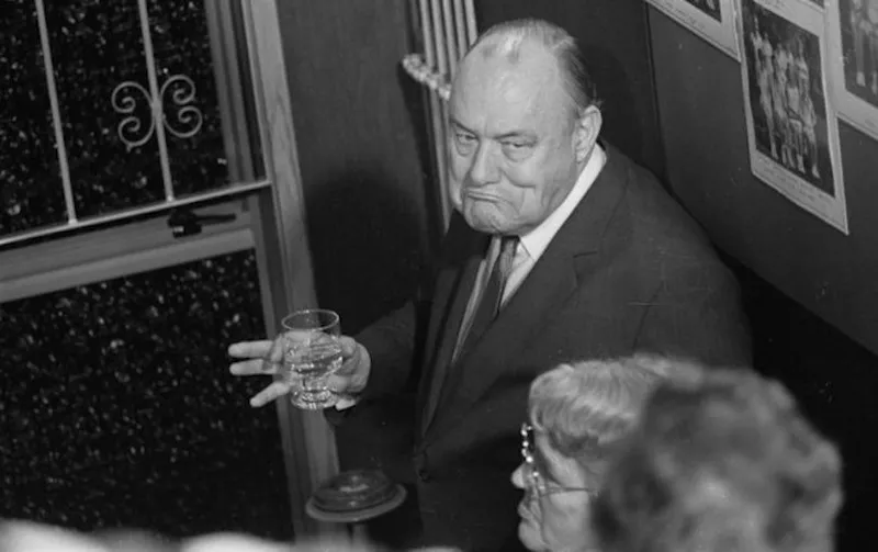 An older man in a suit holds a glass and looks up with a serious or contemplative expression, standing near a window and several people, with framed photos on the wall behind him.