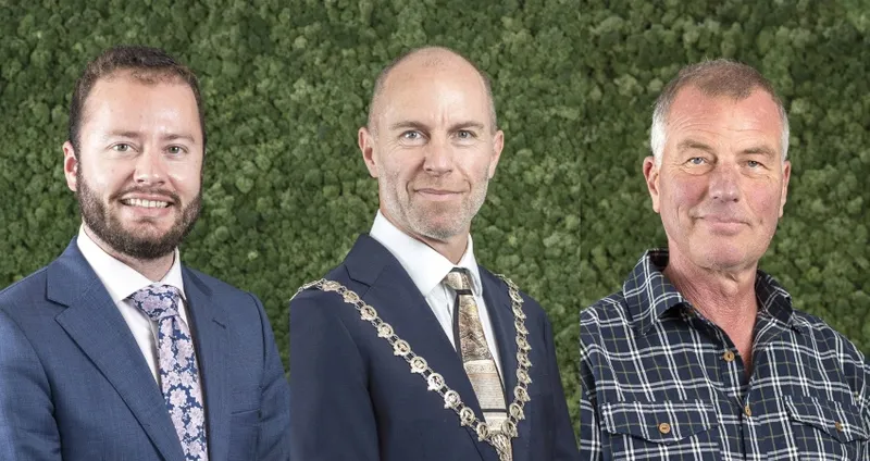 Three men stand in front of a green foliage background