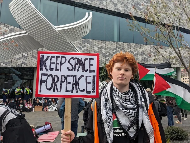 a red haired young man wearing a keffiyeh and holding a sign saying keep space for peace