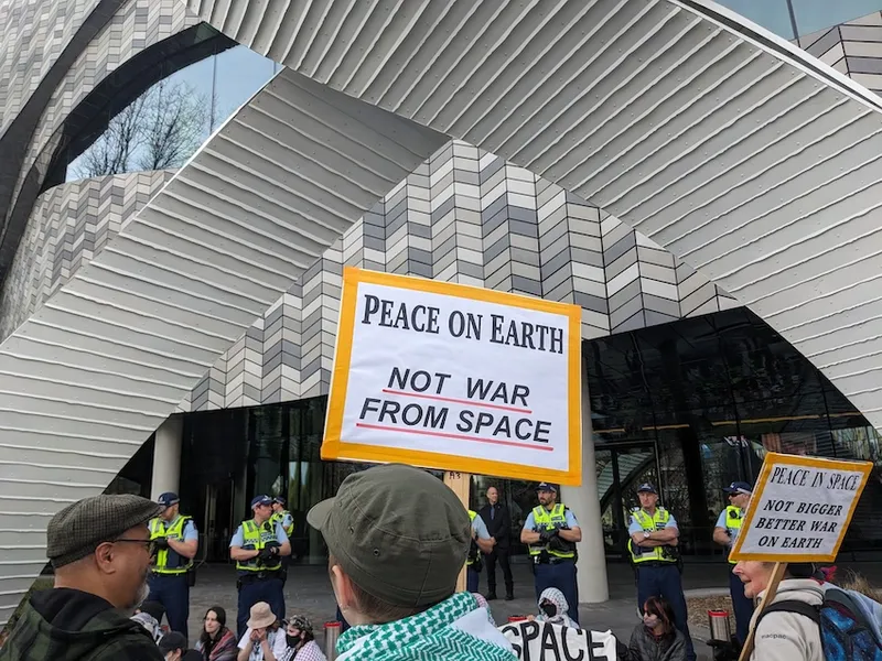 a sign saying peace on earth not war from space, with the grey tiles of Te Pae behind it