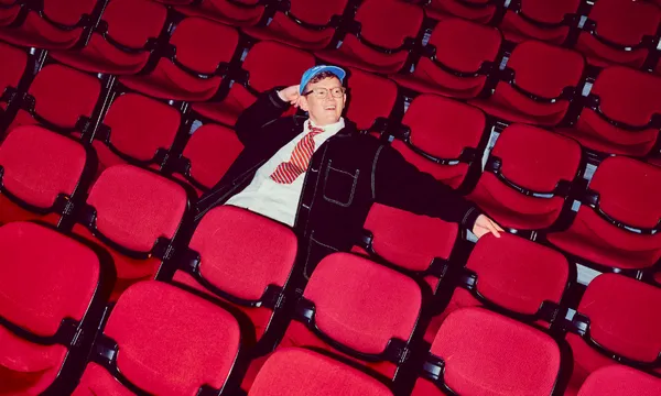 A person in a blue cap and striped tie lounges with one arm behind their head in a nearly empty theater, surrounded by rows of red seats.