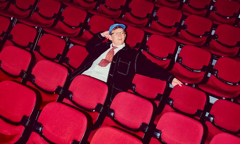 A person in a blue cap and striped tie lounges with one arm behind their head in a nearly empty theater, surrounded by rows of red seats.