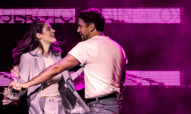 A photograph of the actors in Heartbreak Hotel (a theatre show) on stage which is lit with pink. they are holding hands and are in active poses almost as if they are dancing.