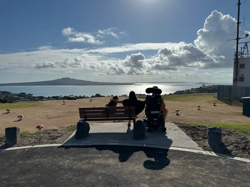a suny day looking over the cone of ranitoto, with a park bench with a person sitting in it and a power wheel chair next to her
