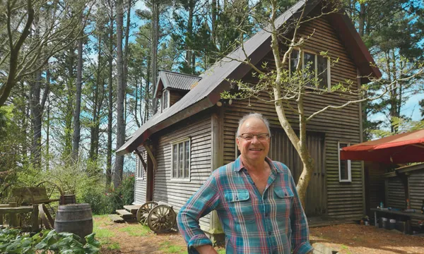 A smiling older man in a plaid shirt stands in front of a rustic wooden house surrounded by trees and greenery on a sunny day.