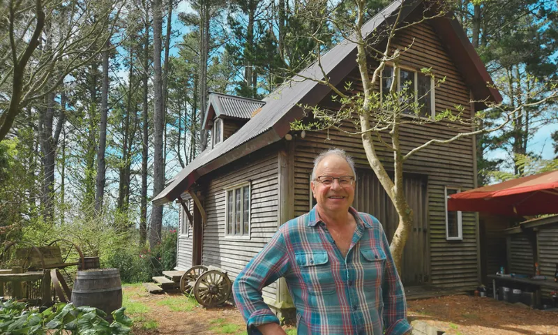 A smiling older man in a plaid shirt stands in front of a rustic wooden house surrounded by trees and greenery on a sunny day.