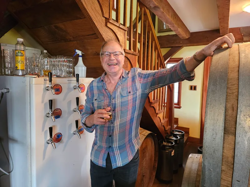 A smiling man in a plaid shirt holds a drink and leans against a large barrel in a rustic room with wooden beams, a staircase, and a row of beer taps attached to a fridge. Glasses and bottles are on top of the fridge.