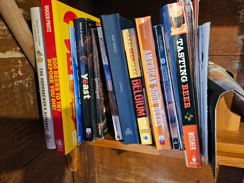 A collection of books about beer and brewing, including titles such as "yeast," "Beer Guide Belgium," "American Sour Beers," and "Tasting Beer," arranged on a wooden shelf.