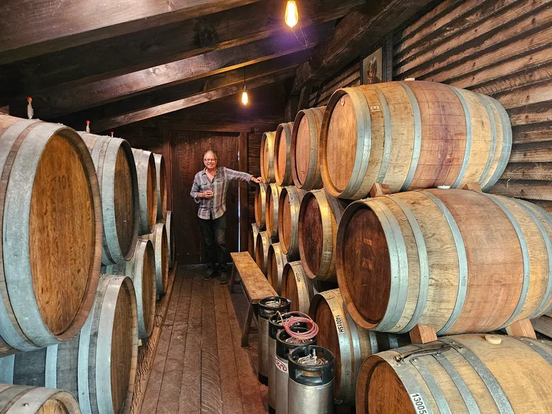 A person stands smiling in a rustic wooden room lined with large stacked barrels on both sides, holding a wine thief, with dim lights hanging from the ceiling.