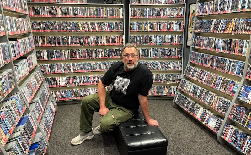 A man sits in the middle of a room surrounded by hundreds of DVDs