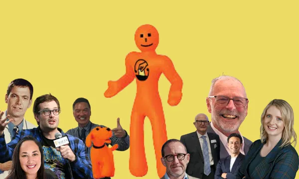 A group of smiling people pose around a large, orange, cartoon figure and a small orange dog, both set against a plain yellow background. The cartoon figure has a simple face and logo on its chest.