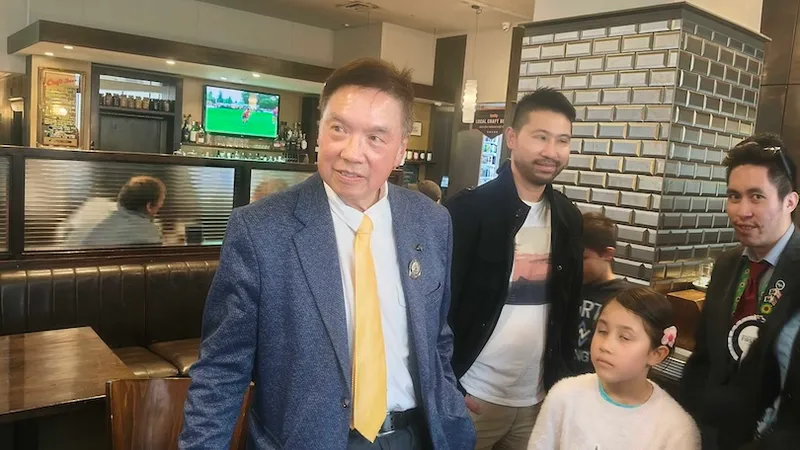 A man in a blue jacket and yellow tie stands in a restaurant with three other people, including a young girl. Behind them, a TV shows a sports game. The setting appears casual and lively.