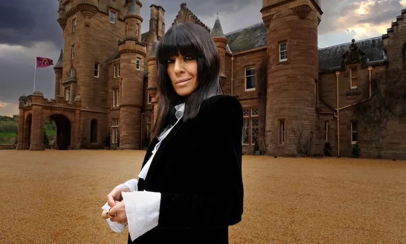 TV presenter Claudia Winkleman wears a black jacket with white cuffs and stands in front of a Scottish castle and looks directly at the camera