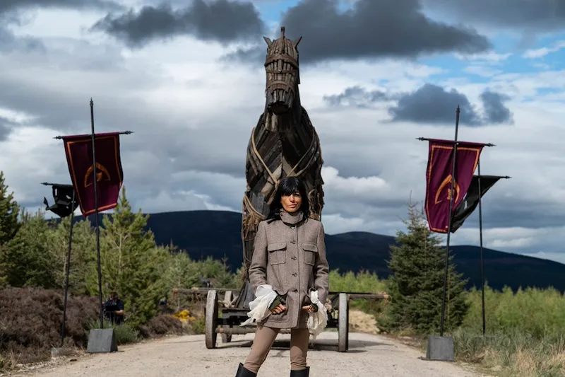 Claudia Winkleman stands on a gravel road with her hands in her pockets. Behindd her is a large Trojan horse and stormy skies