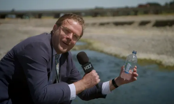 a man in a suit and holding a microphone crouches down next to a stream and holds up a plastic bottle of water
