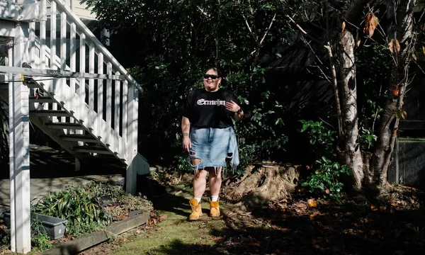 a young woman in a black t-shirt, denim skirt and Timbaland boots stands in her garden wearing dark sunglasses