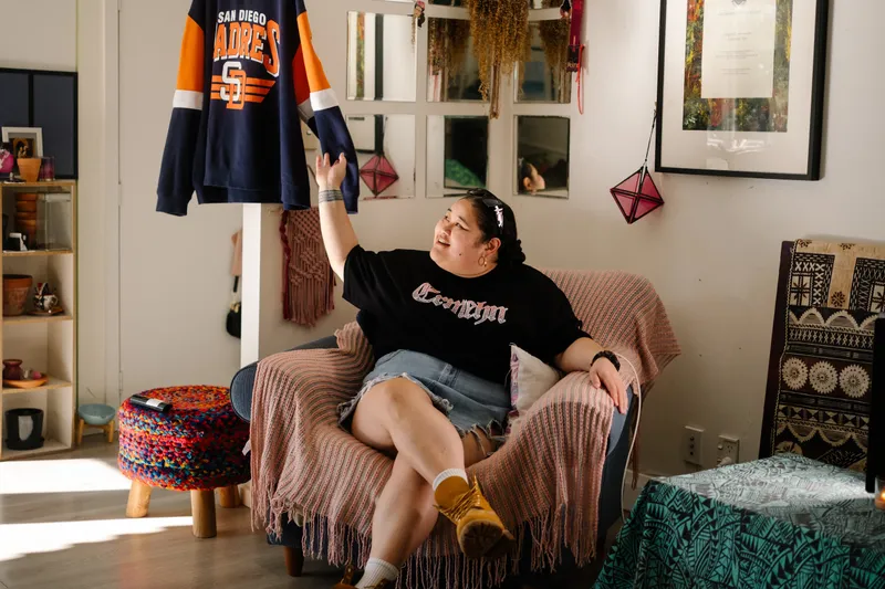 a young woman in a black t-shirt and denim skirt sits in an armchair with her legs crossed. She is gesturing to an American football sweatshirt hanging from the ceiling