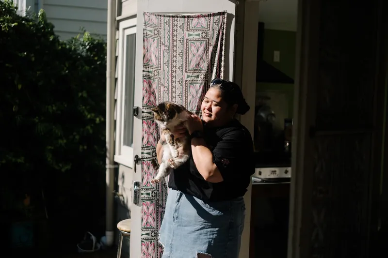 a woman in a black t-shirt and denim skirt stands in the sun holding a white, black and brown cat
