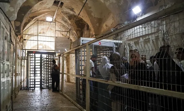 People stand in a crowded, fenced corridor with metal bars, waiting to pass through a security checkpoint with a turnstile gate in an old, worn building with arched ceilings and exposed lights.