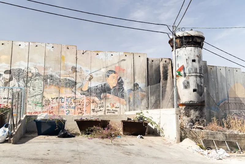 A concrete separation wall with political graffiti, including a figure slinging a stone. A worn watchtower stands to the right with a Palestinian flag. Trash bins and cables are visible in the foreground under a clear sky.