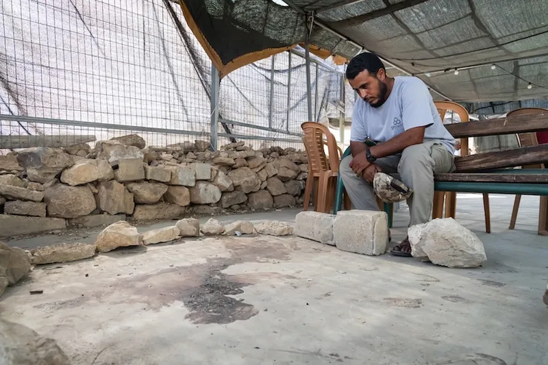 A man sits on a bench under a canopy, observing the ground, with stone walls and plastic chairs in the background.