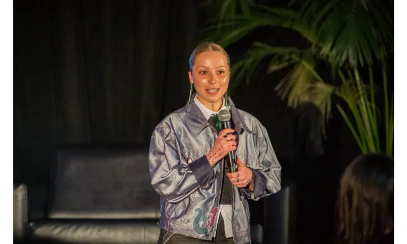 A young woman holds a microphone and is speaking into it. She has a shiny jacket on and pulled back hair.