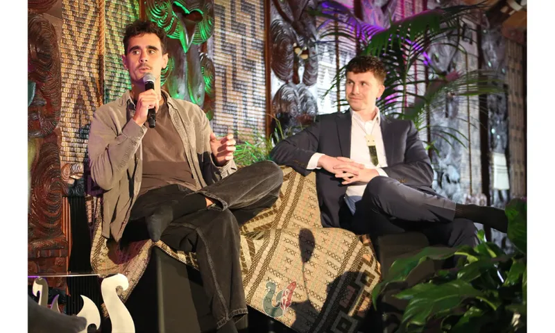Two young men sit on a stage at a marae. The man on the left has a microphone and is talking.