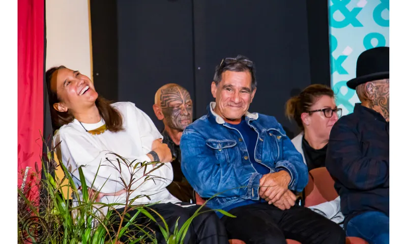 Five people seated on a stage and smiling. One woman has her head thrown back, laughing. They are all Māori.