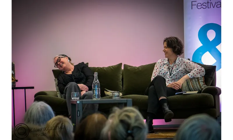 Two women are seated on a couch. The woman on the left is leaning back laughing. The woman on the right is looking at her and smiling.