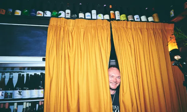 A smiling man peeks out from behind yellow curtains in a bar with shelves of wine bottles above and behind him. The lighting is warm and the atmosphere appears cozy and inviting.