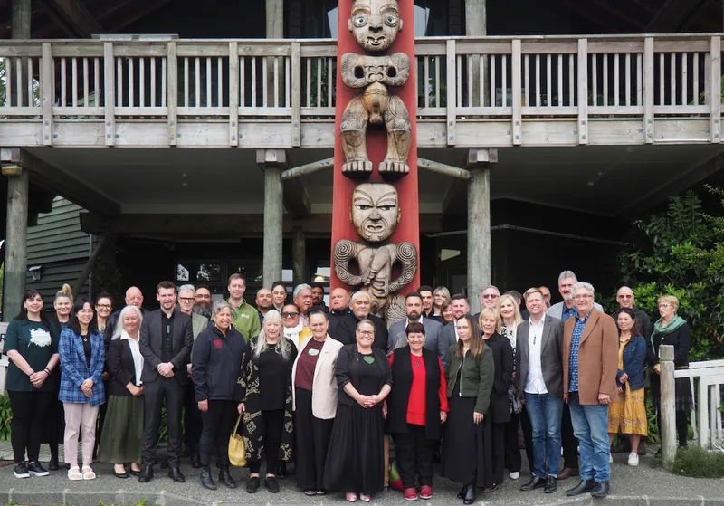 A large group of people stand in front of a carved wooden totem pole and a traditional wooden building, posing for a group photo. The scene is outdoors, with greenery visible on the right.