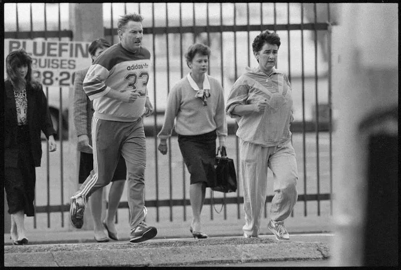 Four adults cross a city street; two wear business attire, while a man and woman in tracksuits jog beside them. A fence and billboard are visible in the background. The image is in black and white.