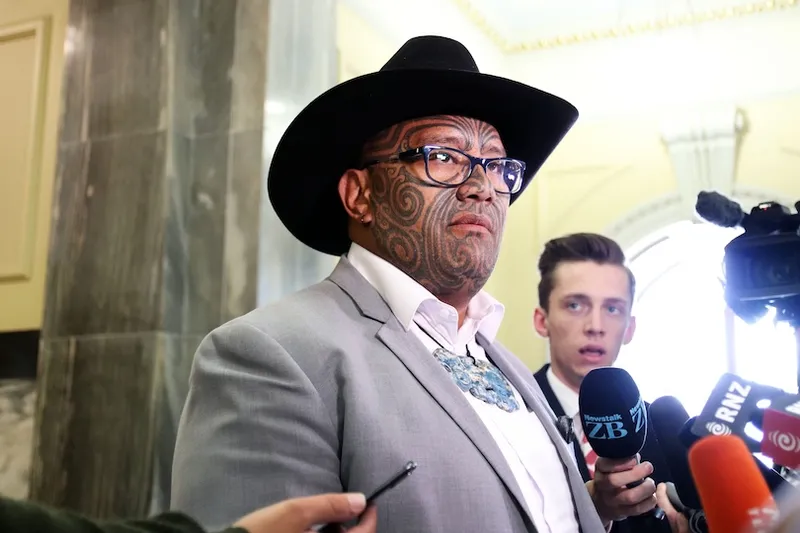 A person wearing a black hat, glasses, and a grey suit with facial tattoos speaks to the media. Several microphones are positioned in front of them, and another person stands nearby.