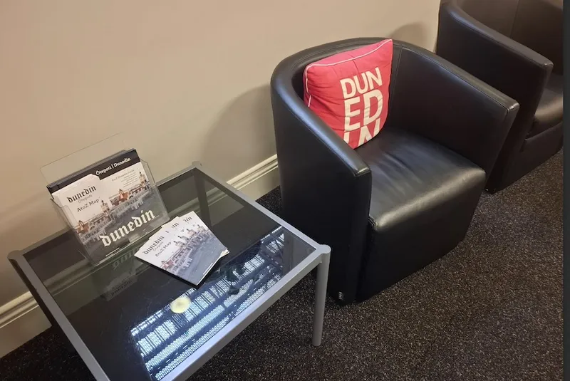 A photo of a seat, a cushion with DUNEDIN written on it, and a coffee table covered in Dunedin maps