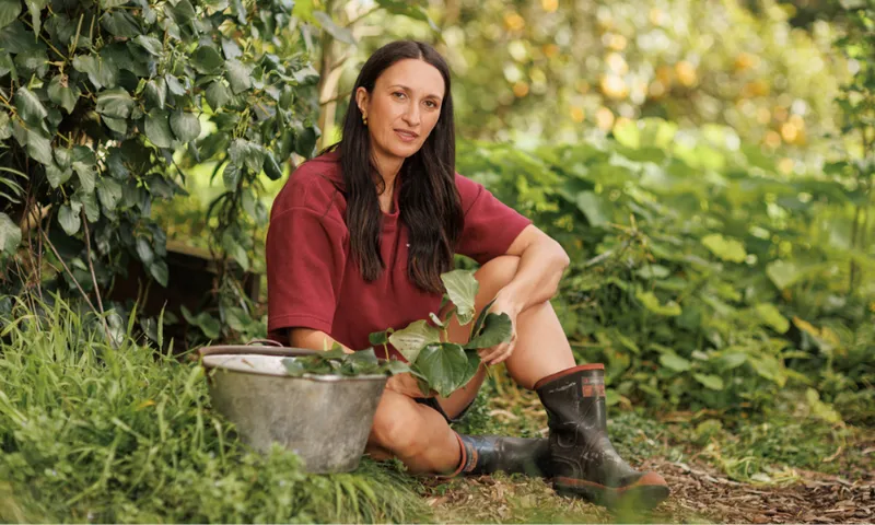A woman with long dark hair, wearing a maroon shirt and black boots, sits on the ground in a lush garden, holding green leaves with a metal bucket of harvested greens beside her.