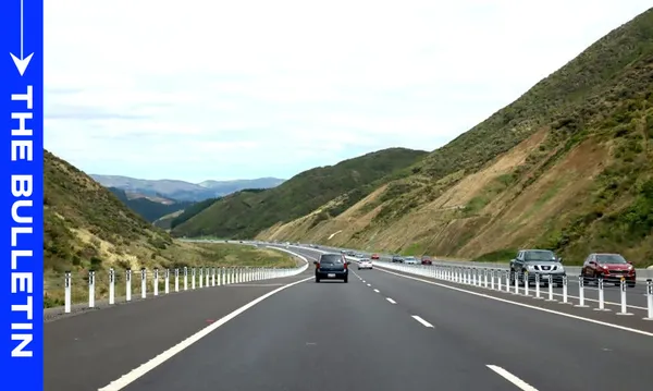 Cars drive along a multi-lane highway winding through green hills under a cloudy sky, with a blue vertical banner on the left reading “THE BULLETIN.”.