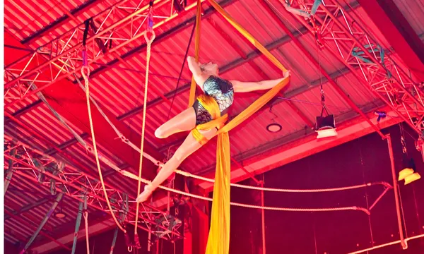A performer in a leotard gracefully poses on yellow aerial silks, suspended high above the ground in a brightly lit indoor venue with metal beams and stage lights overhead.