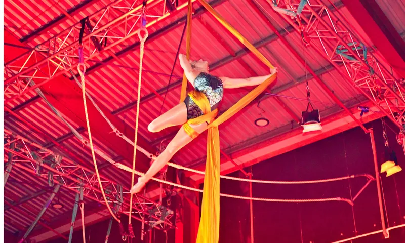 A performer in a leotard gracefully poses on yellow aerial silks, suspended high above the ground in a brightly lit indoor venue with metal beams and stage lights overhead.