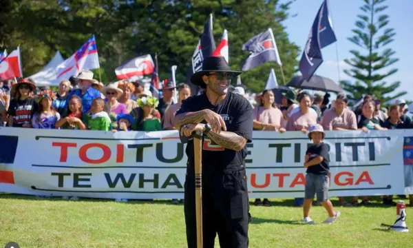 A man in black stands in front of a large crowd holding a banner that reads "TOITŪ TE TIRITI ME TE WHAKAPUTANGA." People in the crowd wave flags and wear hats and sunglasses on a sunny day with trees in the background.